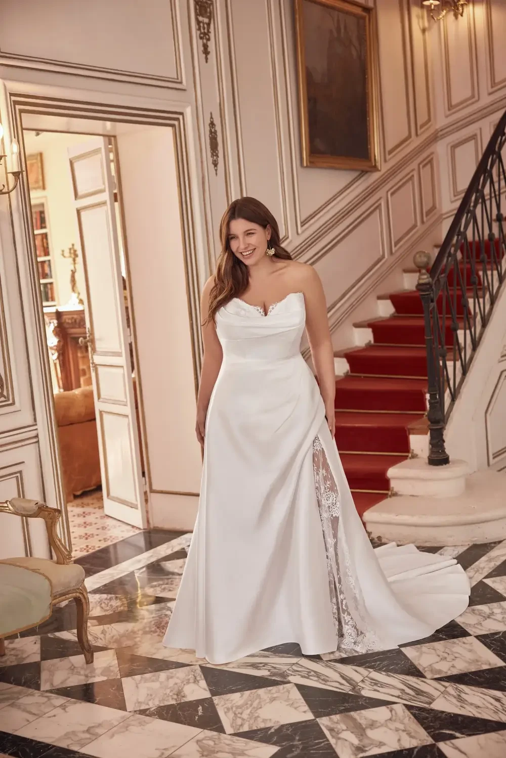 A bride in a strapless white wedding gown with a side slit, standing on a marble floor in an elegant interior with a staircase.
